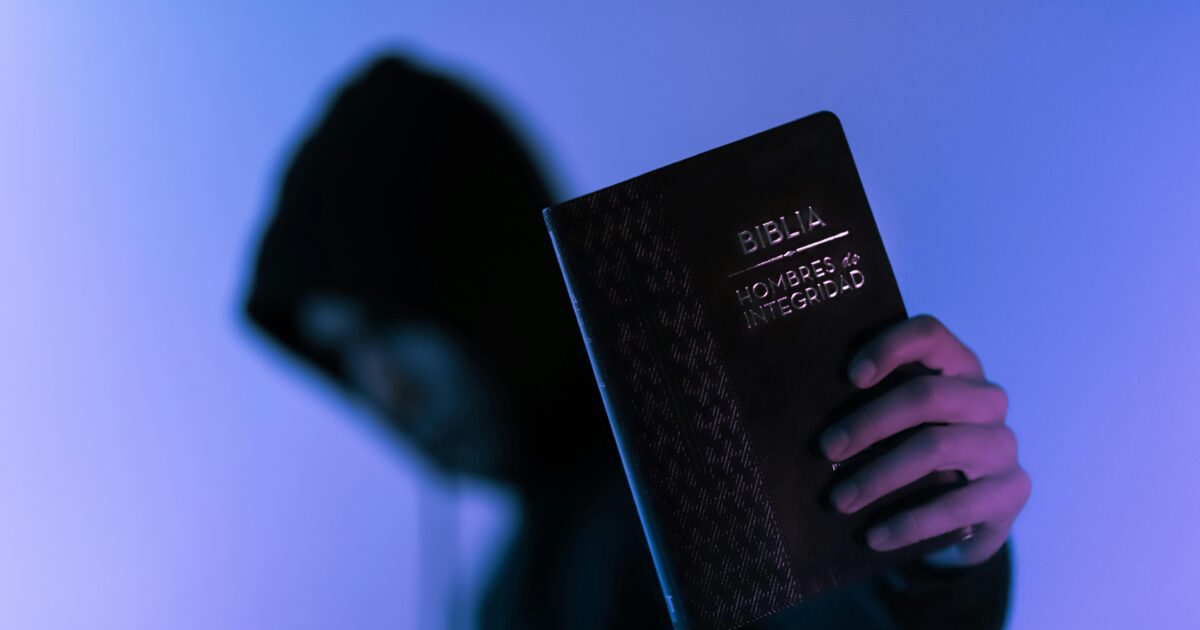 A hooded figure holds a Bible in low-light setting, emphasizing spirituality and mystery.