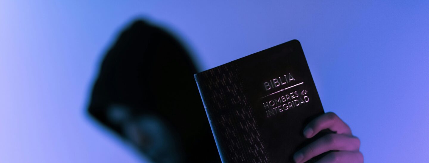 A hooded figure holds a Bible in low-light setting, emphasizing spirituality and mystery.