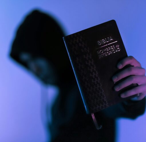 A hooded figure holds a Bible in low-light setting, emphasizing spirituality and mystery.