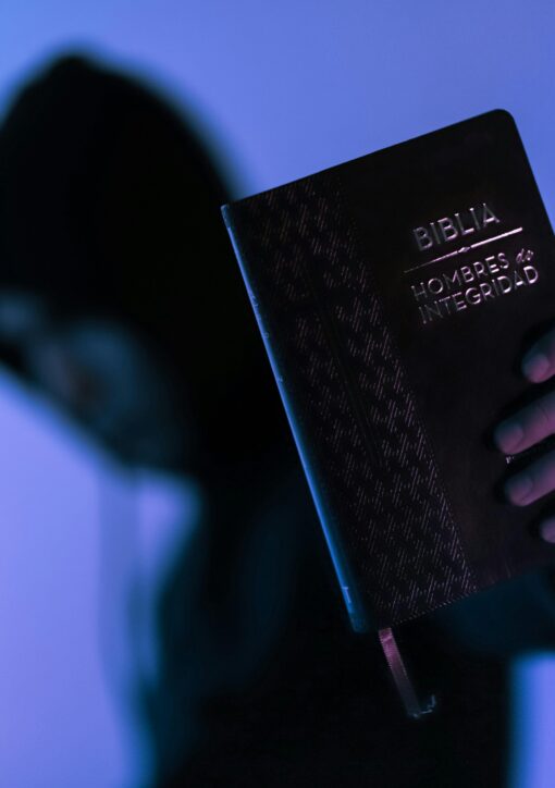 A hooded figure holds a Bible in low-light setting, emphasizing spirituality and mystery.