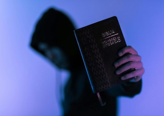 A hooded figure holds a Bible in low-light setting, emphasizing spirituality and mystery.