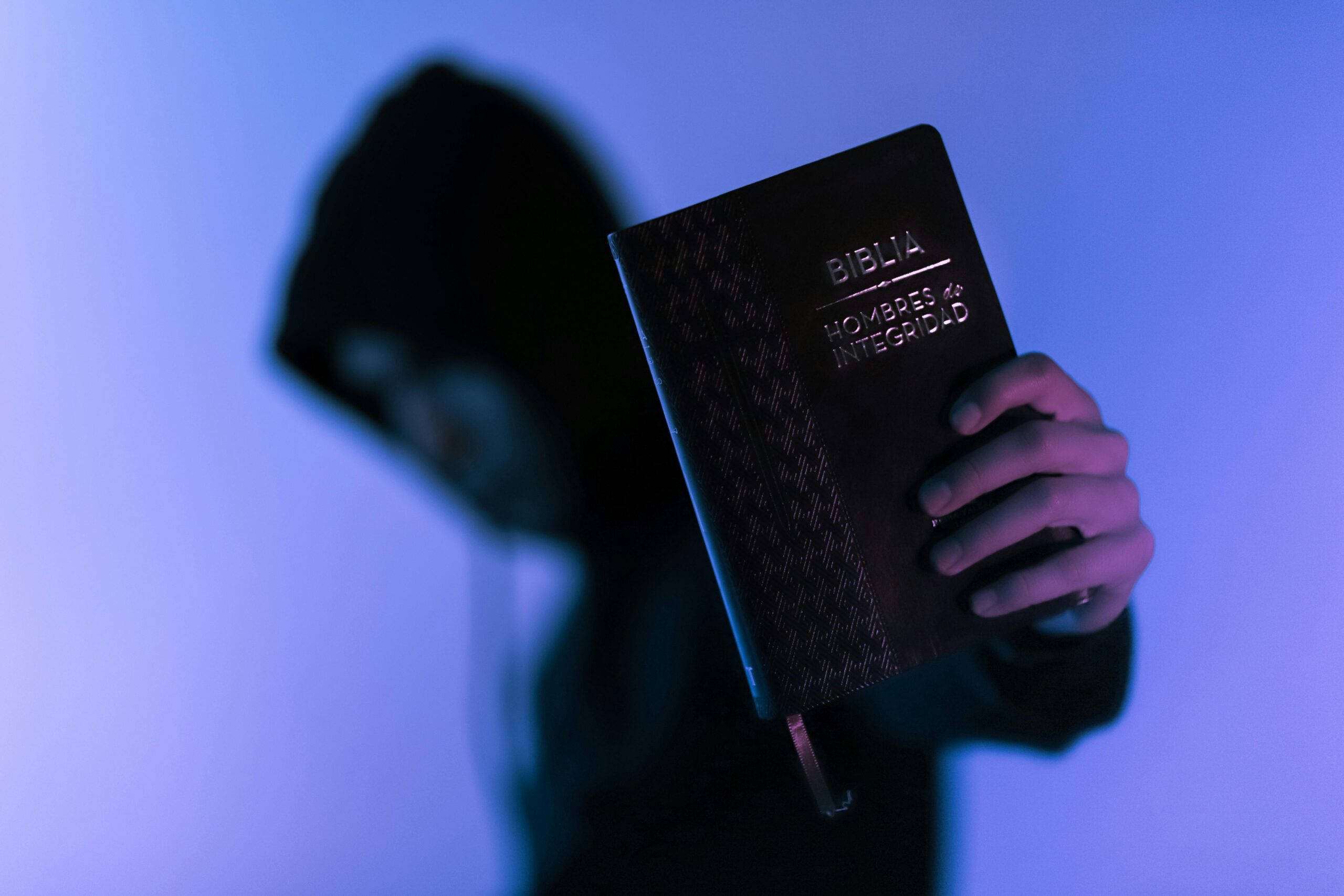 A hooded figure holds a Bible in low-light setting, emphasizing spirituality and mystery.